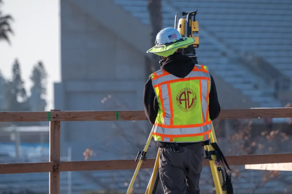 Man wearing safety vest and helmet with surveying equipment outdoors.