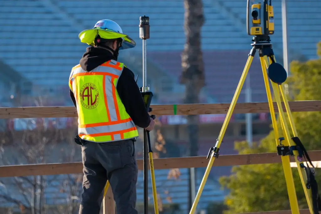 Man wearing safety vest and helmet with surveying equipment outdoors.