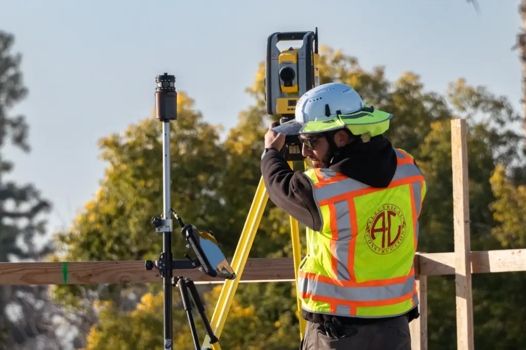 Man wearing safety vest and helmet with surveying equipment outdoors.