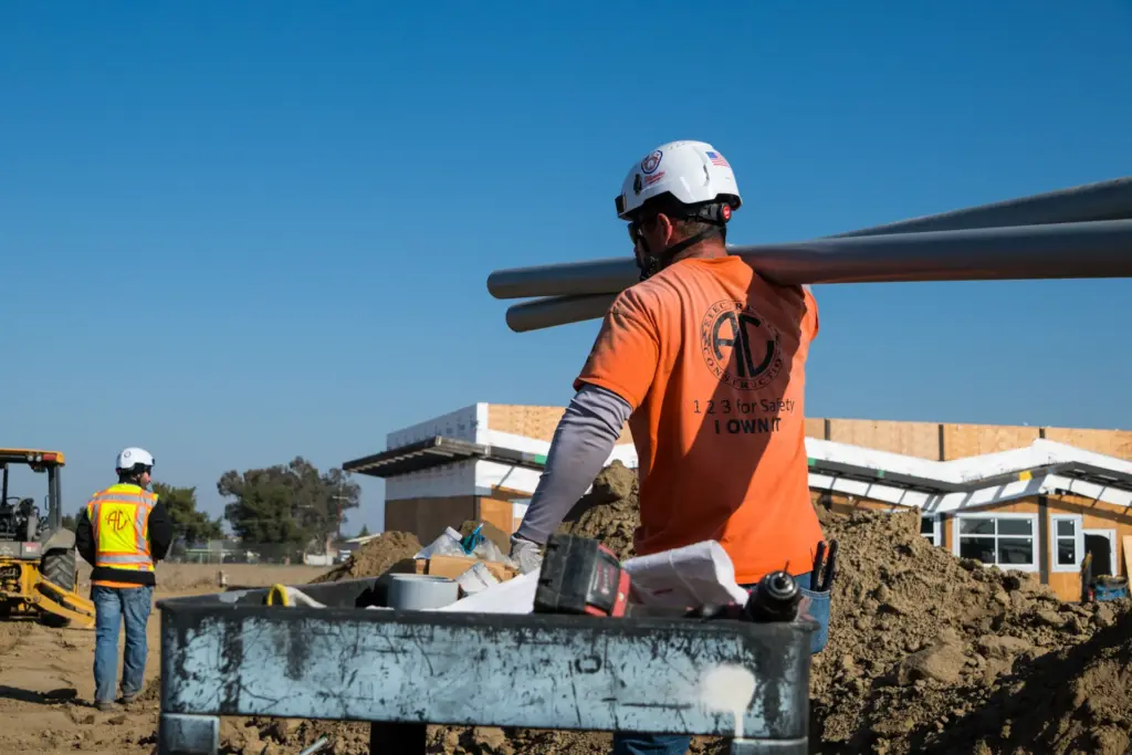 A construction worker is carrying large conduit pipes while another works nearby.