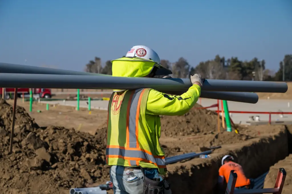 A construction worker is carrying large conduit pipes while another works nearby.