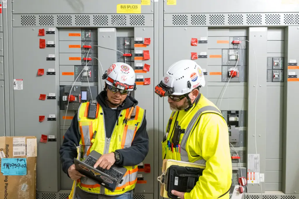 Two construction workers wearing safety gear and helmets stand before an electrical control panel with various switches and buttons.