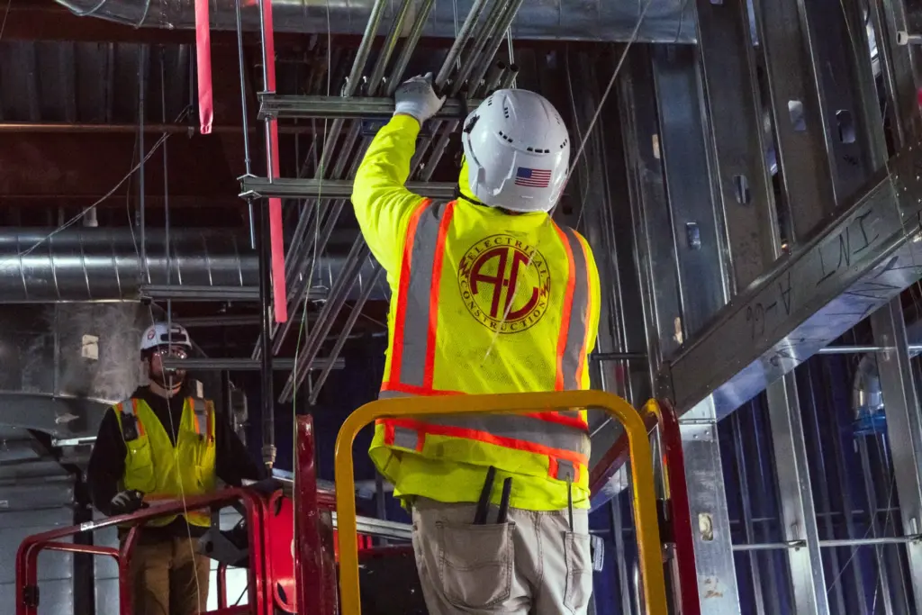 Two electrical construction workers wearing safety gear and helmets are working at a high platform with scaffolding.