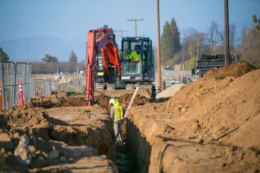 A construction worker is digging a trench with an excavator nearby.
