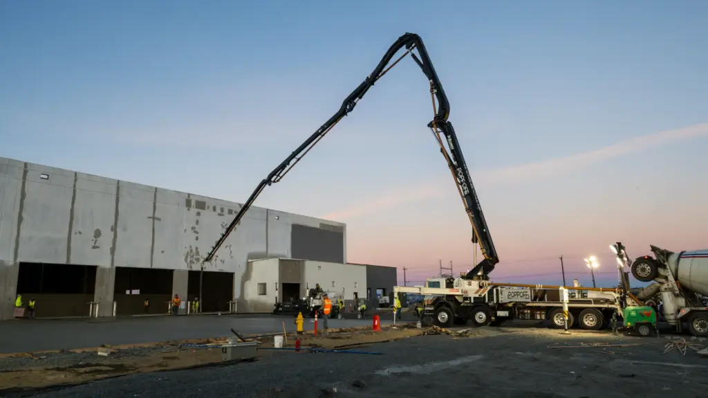 A large concrete pump truck is parked next to a building under construction at sunrise.