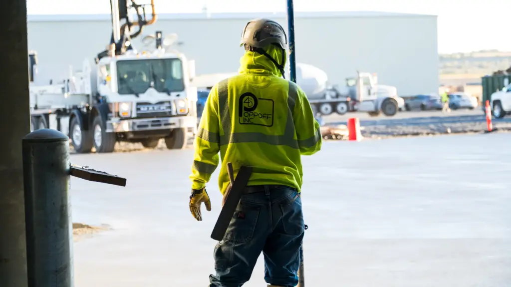 A construction worker is walking away from a truck and building site with his back to us.