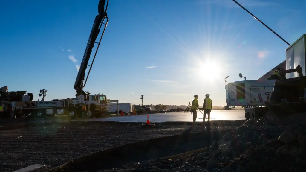 Two workers stand near a concrete pump under bright sunlight with buildings and sky in background.