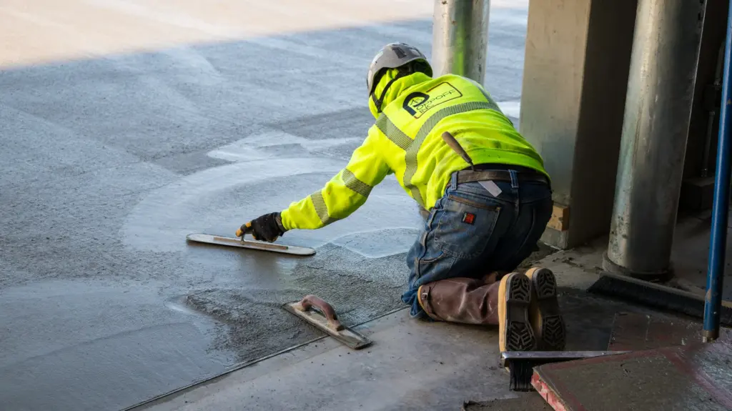 A construction worker is laying down concrete with a trowel and other tools near him.