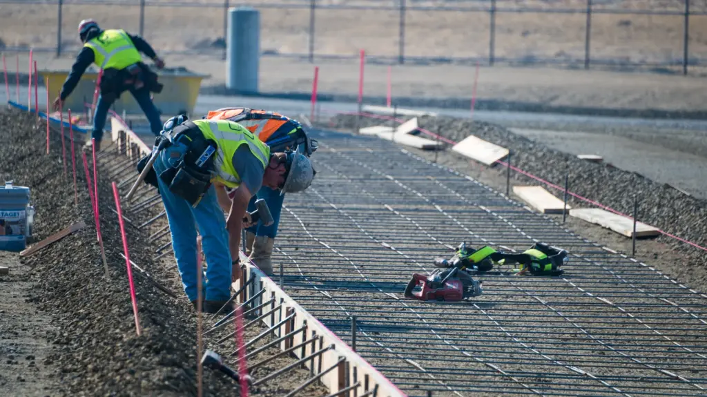 Two construction workers are working with tools to secure a rebar support layer for a concrete slab.