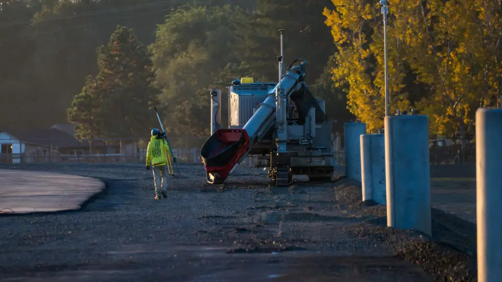A person walking with a surveying tool toward a concrete curb machine.