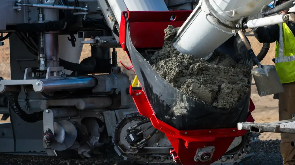 A person is loading concrete from a mixer truck into a curb machine hopper.