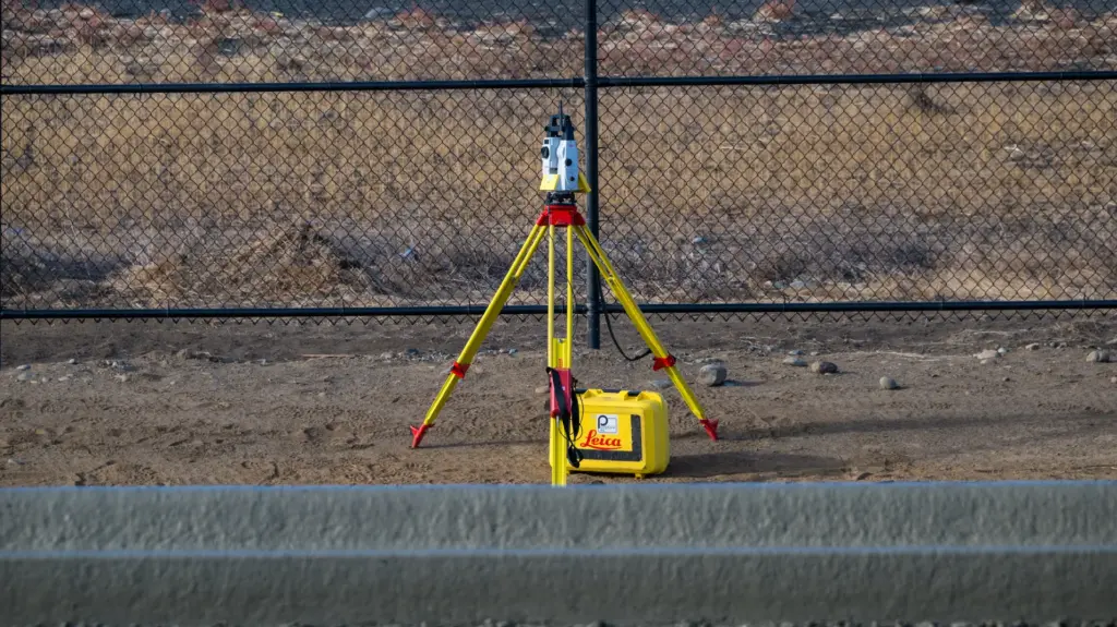 A yellow tripod with a total machine is set up against a chain link fence.