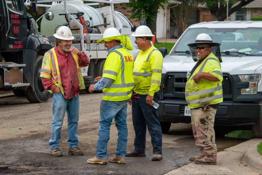 Four construction workers wearing hard hats and safety vests stand near a white truck.