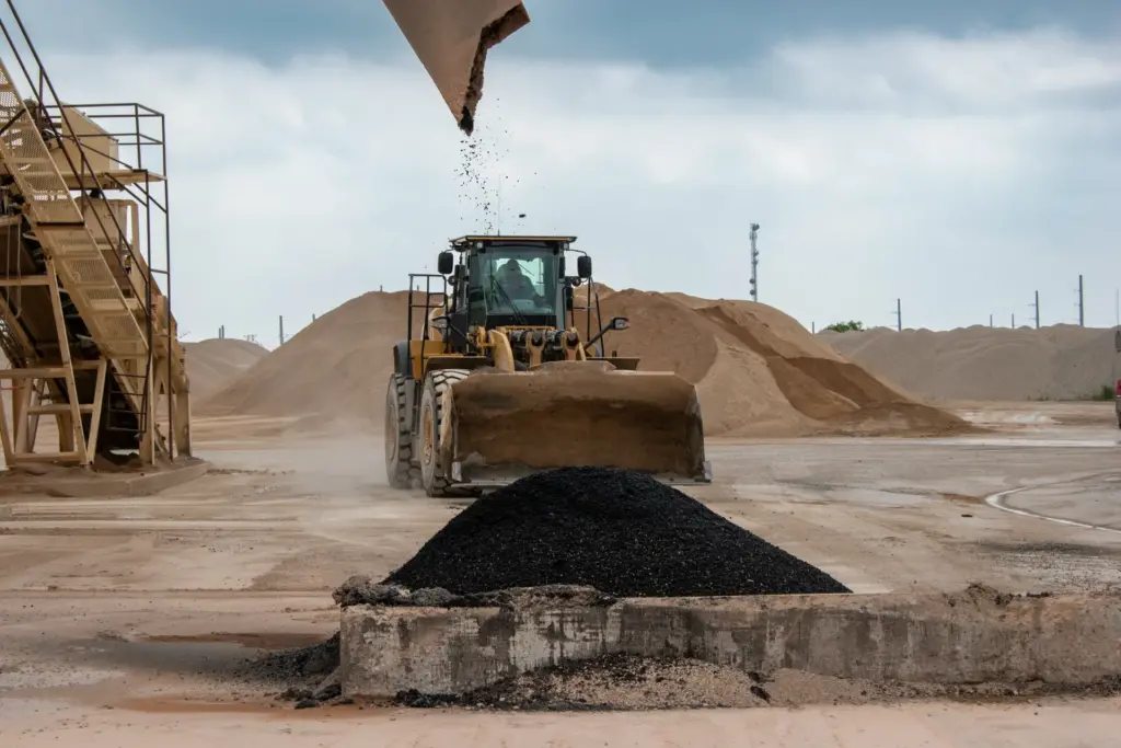 A bulldozer is working at an asphalt plant near a pile of asphalt.