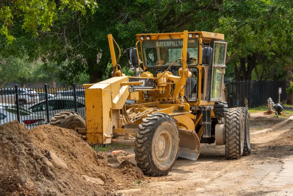 A yellow road scraping machine is parked near a dirt pile and trees.