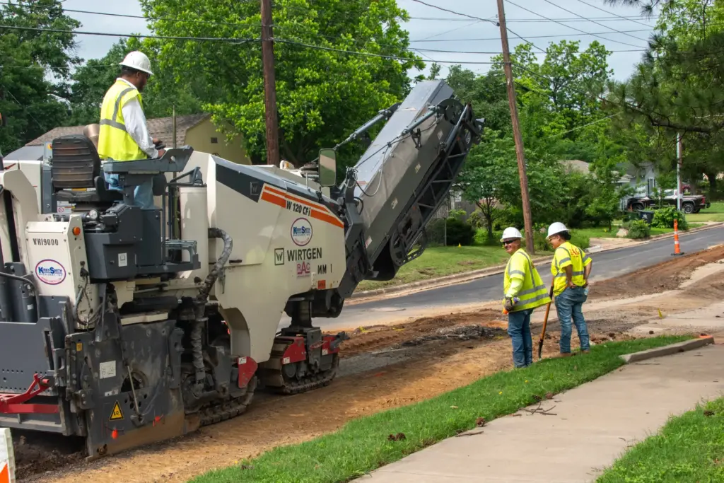 Two construction workers wearing high visibility vests and hard hats stand near a road paving machine.