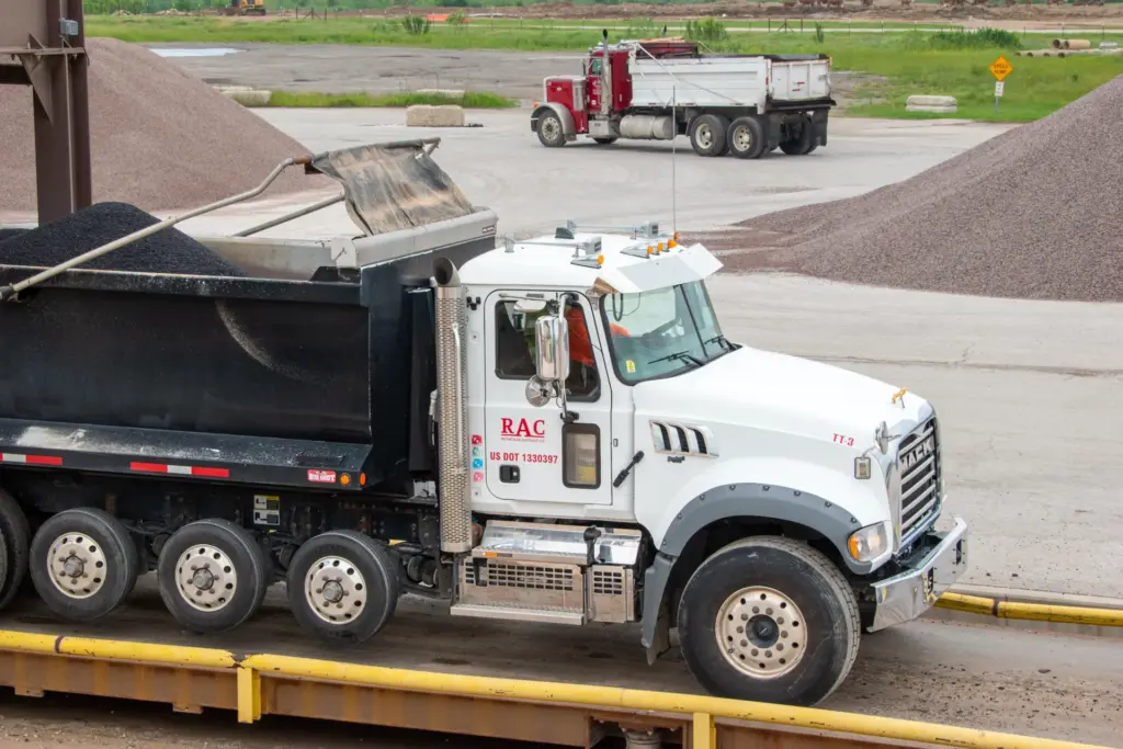 A white truck with a black dump bed drives through an asphalt plant.