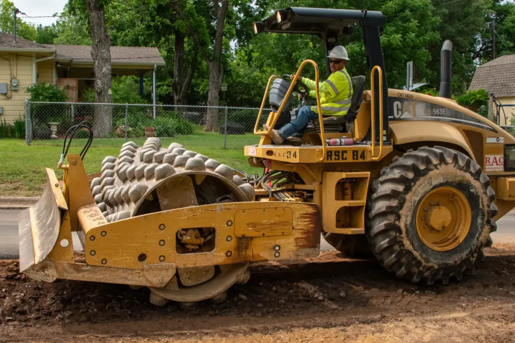 A person is driving a yellow vibratory machine at an excavation site with houses and trees in the background.