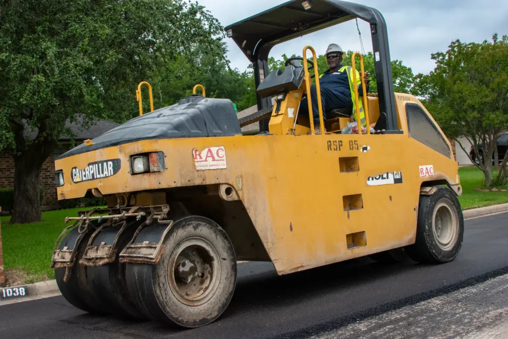A construction worker is driving a road rolling machine amidst trees and machinery.