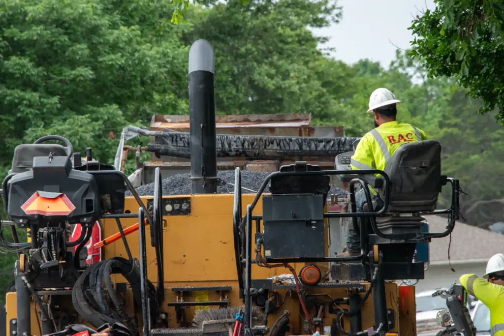 A construction worker is sitting atop a road paving machine amidst trees and machinery.