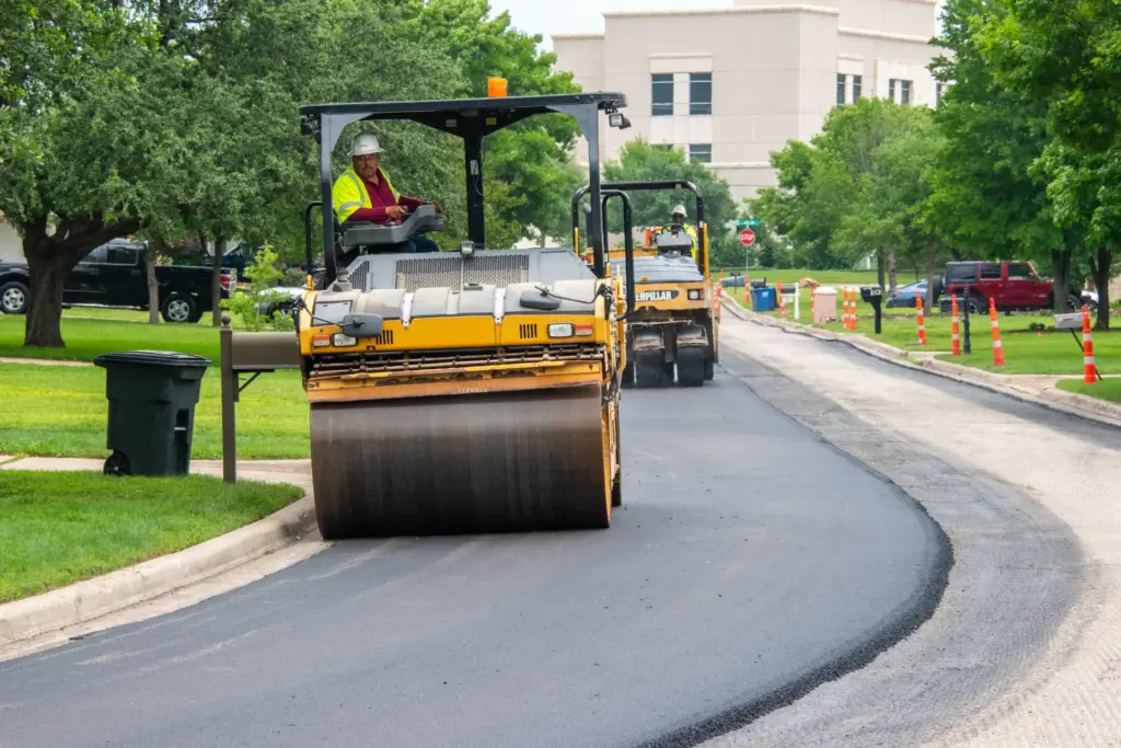A person is driving a yellow road roller down a curved asphalt roadway surrounded by trees and buildings.